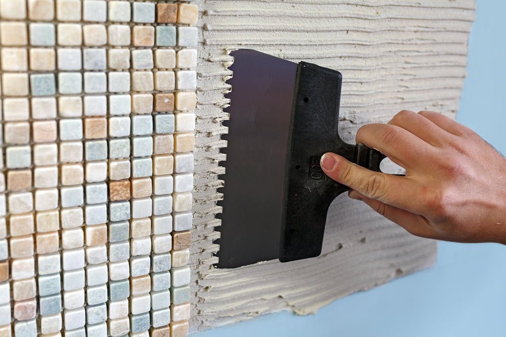 Emmer witte pastalijm met lijmkam naast een vel mozaïektegels, lijm voor mozaïektegels aanbrengen op een wand